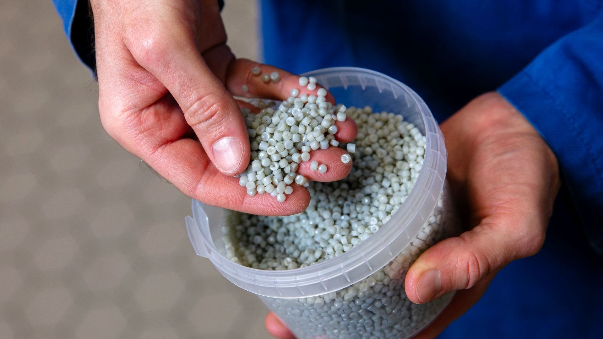 : A close-up of hands holding small, grey plastic pellets. The pellets, which are all uniform in size and shape, are being scooped up from a clear plastic container.