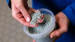 : A close-up of hands holding small, grey plastic pellets. The pellets, which are all uniform in size and shape, are being scooped up from a clear plastic container.