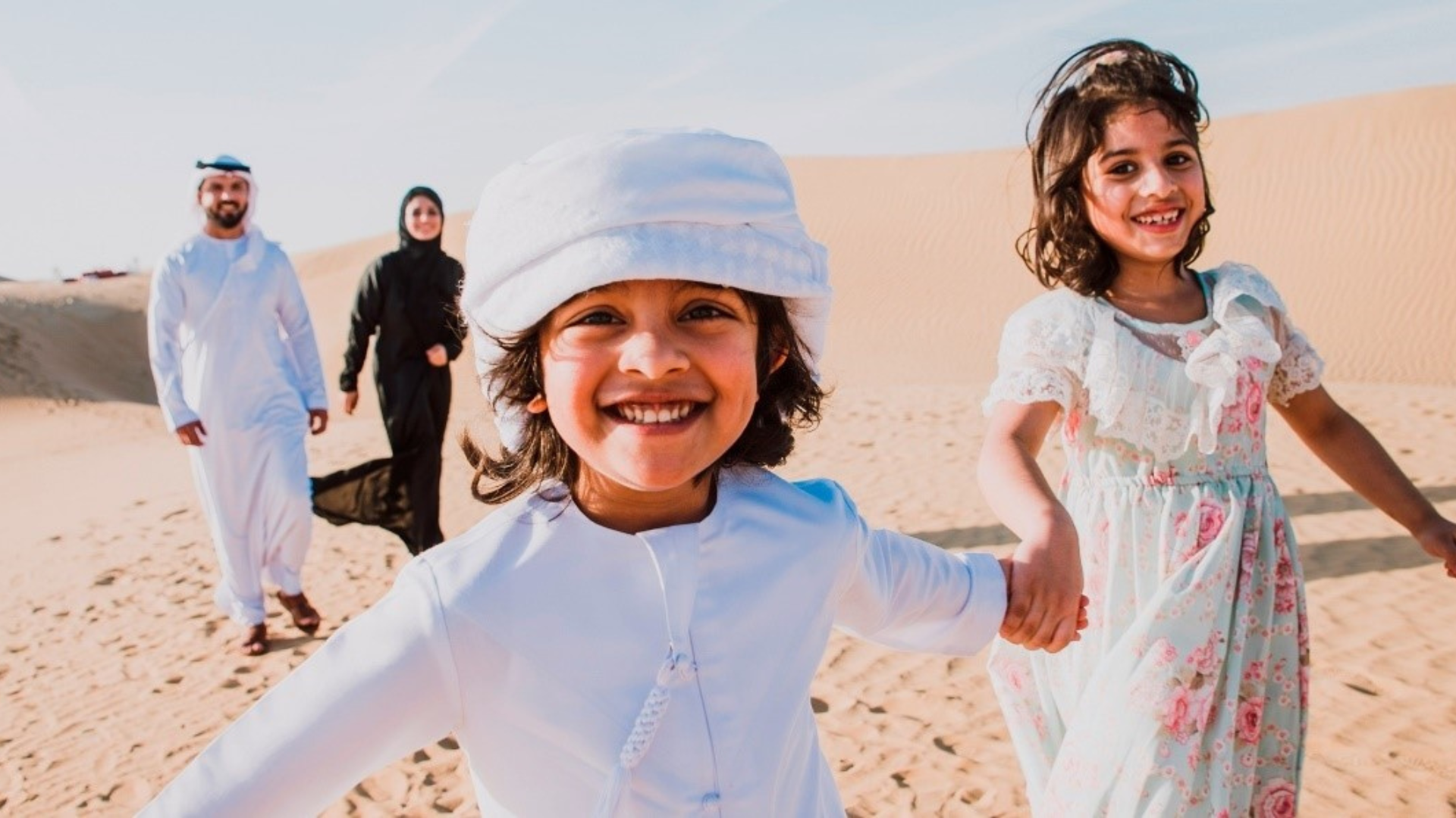 2 kids smiling holding each other's hand and parents at back looking at them