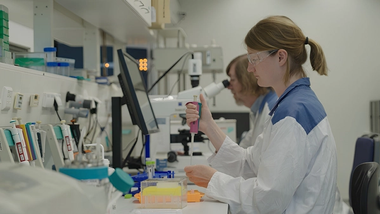 A woman in a lab coat and protective goggles uses a pipette.