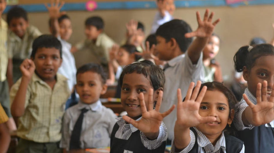 Children in a classroom raise their hands and smile at the camera, having taken part in a Lifebuoy handwashing lesson