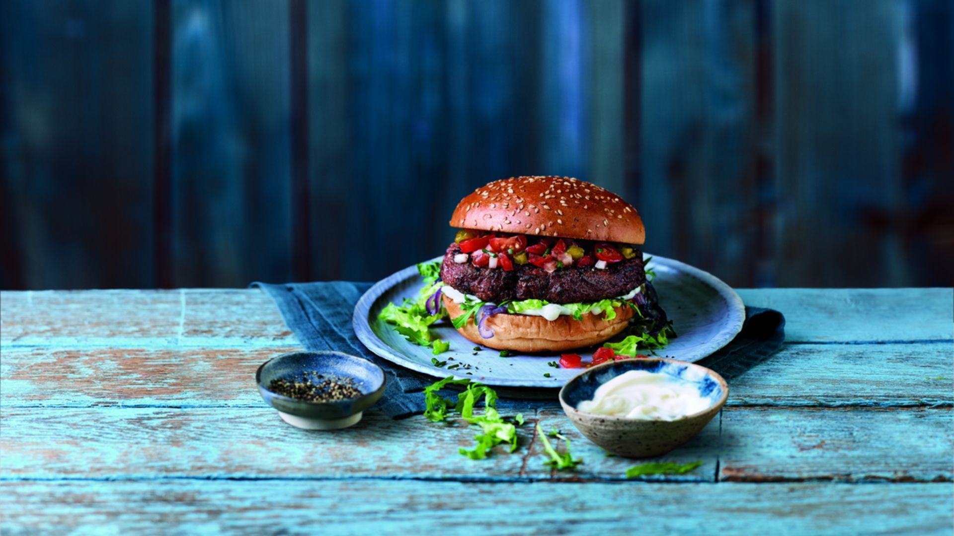 A well garnished burger on a plate, which is on the blue wooden table.