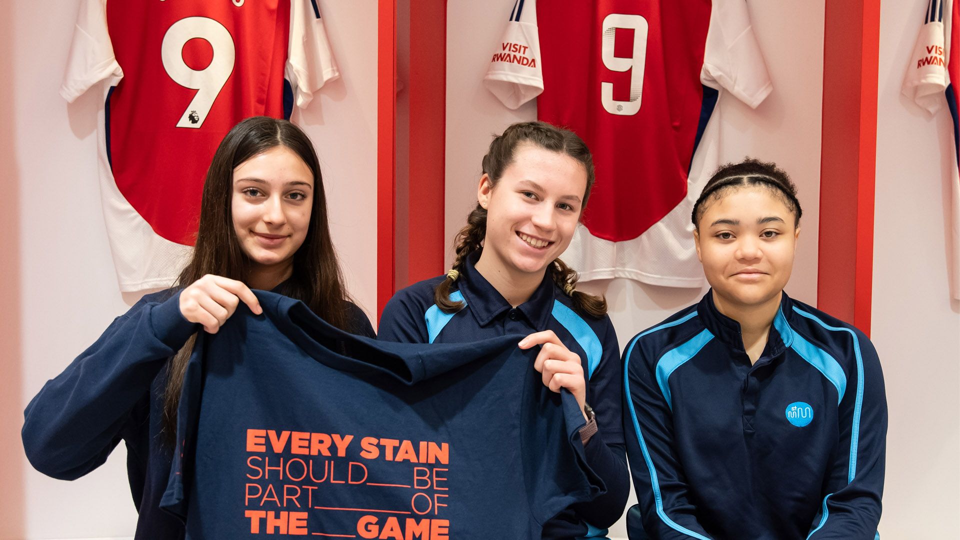 Three girls, sitting in the Arsenal FC changing room, holding a t-shirt that reads “Every stain should be part of the game”.