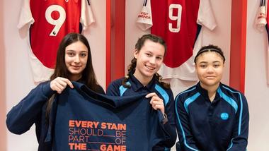 Three girls, sitting in the Arsenal FC changing room, holding a t-shirt that reads “Every stain should be part of the game”.