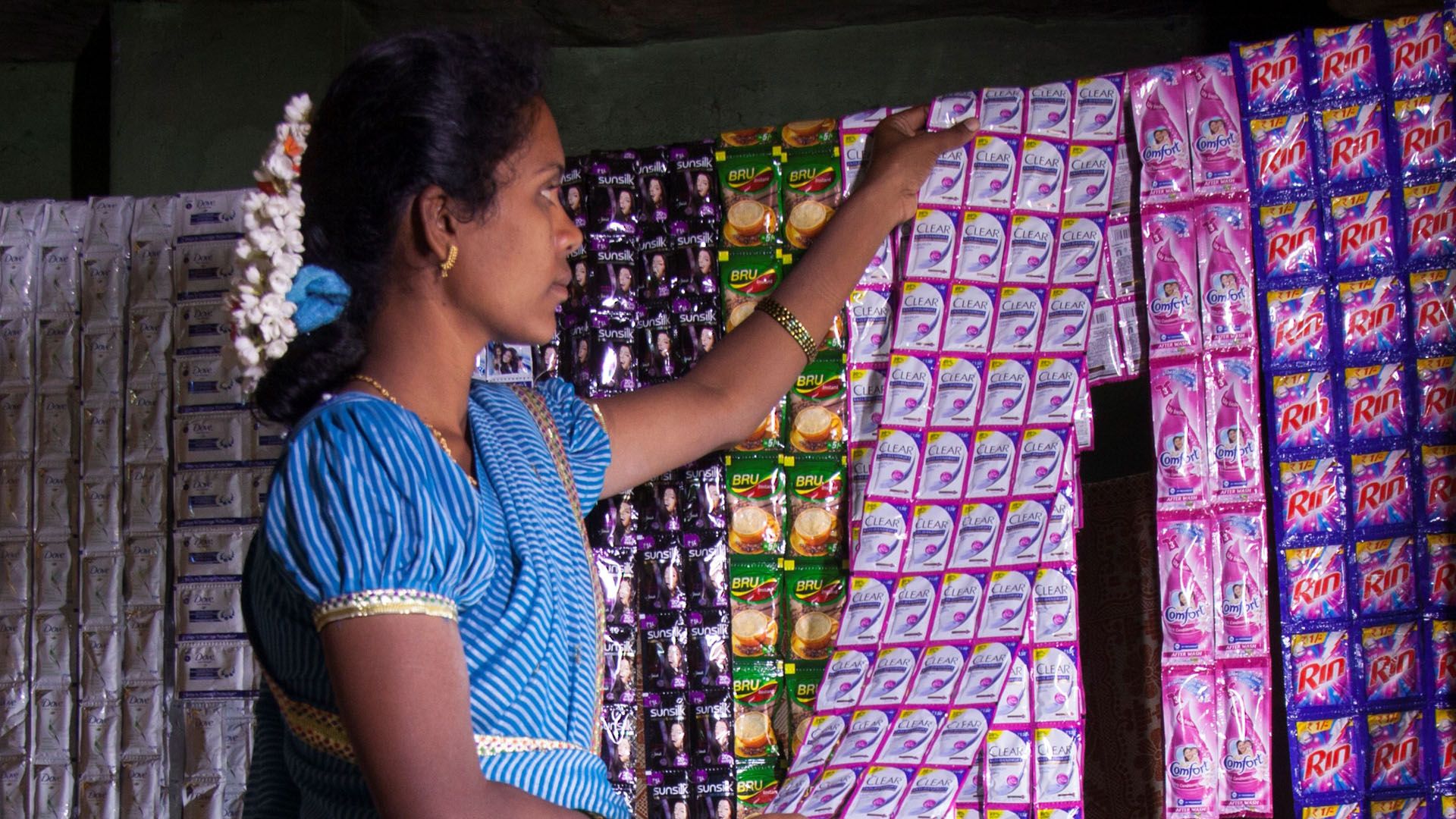 Woman store owner hanging up a sheet of sachets of Clear shampoo. Also visible are sachets of Rin, Bru, Sunsilk and Comfort.